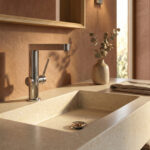 Minimalist bathroom design featuring the Finoris 230 basin mixer on a beige Corian countertop with a classic chrome drain and wooden interior accents under natural sunlight
