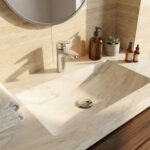 A close-up shot of a modern, integrated Corian sink and countertop with a light beige marble pattern, designed using the KORTE.design 3D configurator. The sink features a hansgrohe Logis 100 chrome single-lever faucet and a matching round drain cover. The vanity is made of dark, rich wood, set against warm-toned marble wall tiles. Soft sunlight casts elegant shadows across the surface, which is decorated with amber glass bottles, a small succulent in a concrete pot, and neatly folded white towels