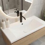A bright, minimalist bathroom with a light speckled Glacier White Corian countertop and integrated sink. The design features a matte black faucet, a round backlit mirror, and a wooden shelf, creating a clean and airy atmosphere