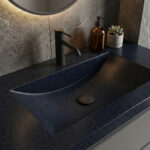 A detailed close-up of a Deep Night Sky Corian countertop featuring an integrated ramp-style sink with a matte black faucet. The dark, starry-textured surface is elegantly styled with a small succulent and an amber soap bottle under soft, warm lighting