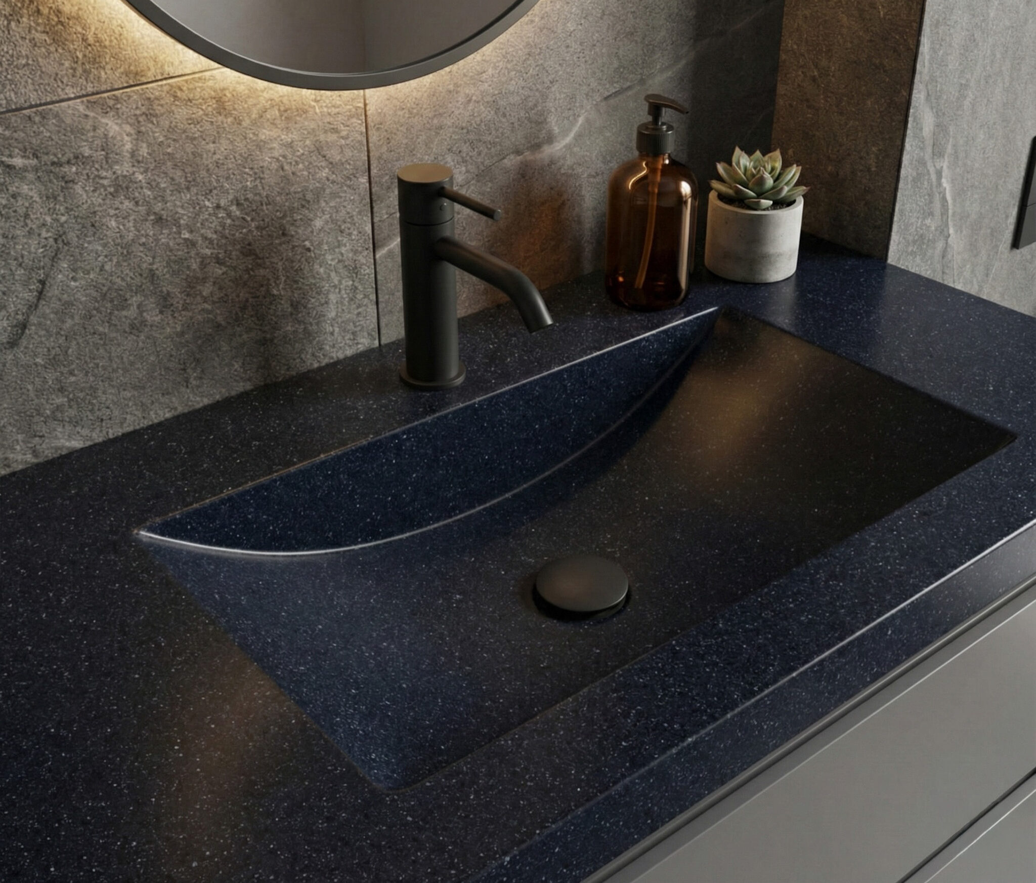 A detailed close-up of a Deep Night Sky Corian countertop featuring an integrated ramp-style sink with a matte black faucet. The dark, starry-textured surface is elegantly styled with a small succulent and an amber soap bottle under soft, warm lighting