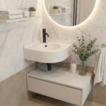 A modern minimalist bathroom corner featuring a curved Glacier White DuPont Corian sink with a matte black faucet. The wall is finished with white marble tiles and a large, round backlit LED mirror. A single white floating shelf on the left holds neatly folded towels and small succulents, while a white floating vanity unit underneath is styled with a soap dispenser, towels, and eucalyptus branches in a glass vase