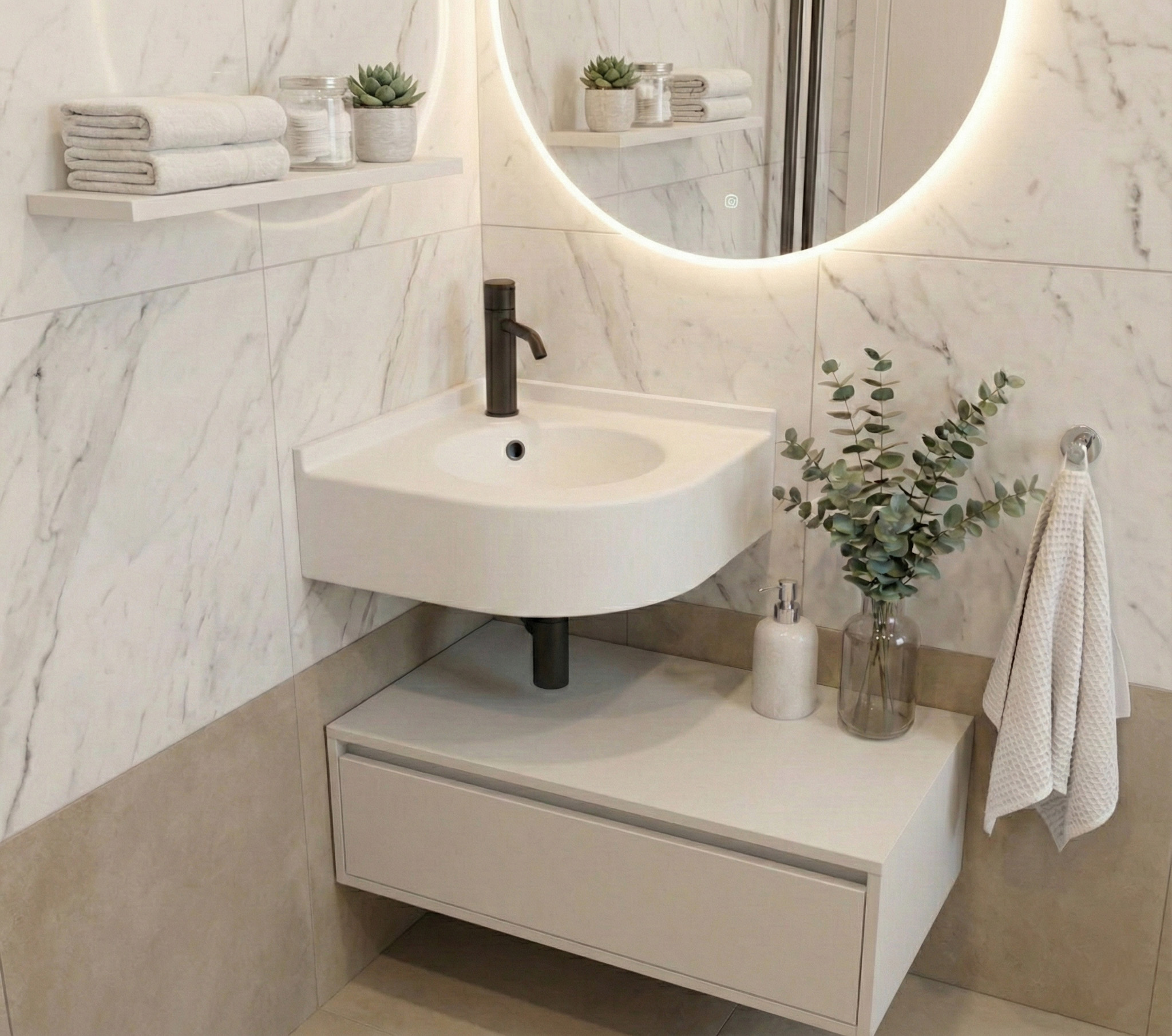 A modern minimalist bathroom corner featuring a curved Glacier White DuPont Corian sink with a matte black faucet. The wall is finished with white marble tiles and a large, round backlit LED mirror. A single white floating shelf on the left holds neatly folded towels and small succulents, while a white floating vanity unit underneath is styled with a soap dispenser, towels, and eucalyptus branches in a glass vase