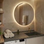 A modern bathroom vanity featuring a grey Corian countertop with an integrated sink and gold faucet. The counter extends over a washing machine and wooden cabinets. Rolled towels, a potted plant, and a gold-framed round mirror are set against beige stone wall tiles