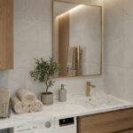 A modern bathroom vanity featuring a white marble-look Corian countertop with an integrated sink and gold faucet. The counter extends over a washing machine and wooden cabinets. Rolled towels, a potted plant, and a gold-framed mirror are set against grey stone wall tiles