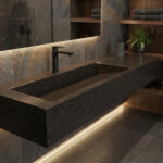 A modern, moody bathroom featuring a long, thick integrated trough sink made of Lava Rock DuPont Corian in a dark charcoal textured finish. The sink is wall-mounted with a matte black faucet, set against dark grey stone tiles with warm LED strip lighting underneath. Dark wood floating shelves with towels are visible in the background