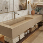 An elegant, warm-toned bathroom with a long integrated trough sink made of Dune Prima DuPont Corian in a sandy-beige finish. Below the vanity is a dark wood floating shelf holding neatly folded towels and wicker baskets. The walls are finished with white marble tiles, decorated with pampas grass and amber glass bottles