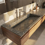 A sunlit bathroom featuring a long, integrated sink and countertop made of Corian in a dark grey terrazzo pattern, designed using the KORTE.design 3D configurator. The floating vanity cabinet below is made of warm walnut wood. A sleek chrome faucet is mounted on the left side of the sink. Strong sunlight streams in from the left, casting long shadows across the Corian surface and the grey hexagonal tile backsplash. Various toiletries, including amber glass bottles and a cream jar, are arranged on the countertop