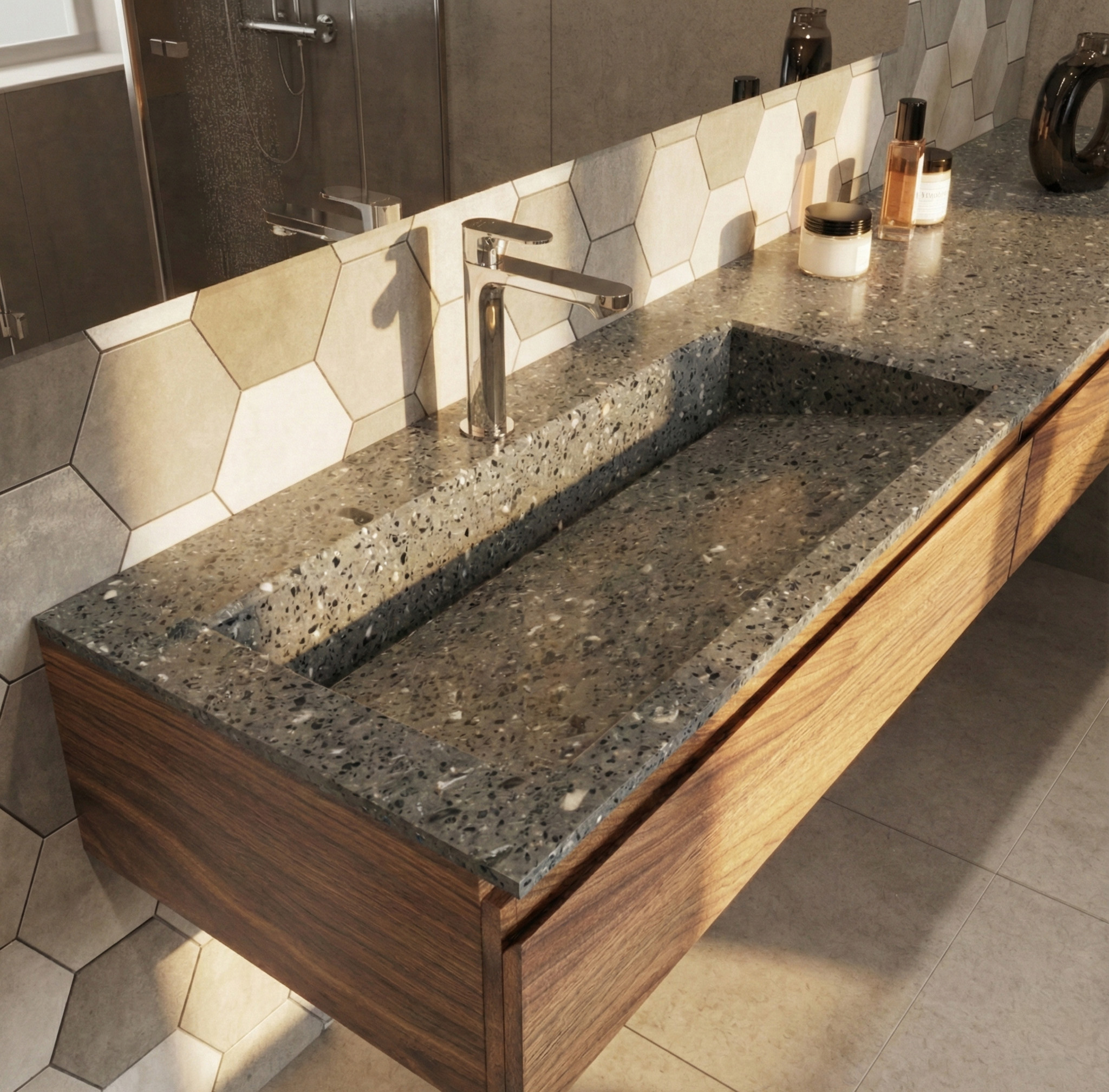 A sunlit bathroom featuring a long, integrated sink and countertop made of Corian in a dark grey terrazzo pattern, designed using the KORTE.design 3D configurator. The floating vanity cabinet below is made of warm walnut wood. A sleek chrome faucet is mounted on the left side of the sink. Strong sunlight streams in from the left, casting long shadows across the Corian surface and the grey hexagonal tile backsplash. Various toiletries, including amber glass bottles and a cream jar, are arranged on the countertop