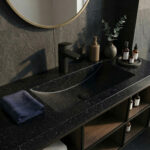 Modern black bathroom Corian Deep Night Sky vanity top with white speckles, a matte black hansgrohe Vivenis 100 faucet, and open wooden shelves.