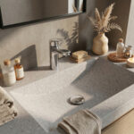 A sun-drenched, detailed close-up of a modern bathroom vanity featuring a seamless Corian solid surface countertop and integrated rectangular sink (speckled pattern with white, grey, and pink chips). A polished Hansgrohe Logis 70 Chrome single-lever basin mixer faucet is the central fixture. The background is a textured grey concrete wall with dramatic leaf shadows cast by natural daylight, a black-framed mirror, and a ceramic vase with dried pampas grass. Beige waffle-texture towels, Patterson brand glass cosmetics bottles, and a bar of soap on a wooden dish are also present.