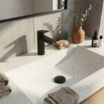 A detailed photograph of a contemporary bathroom vanity countertop. The entire surface is a patterned white Corian solid surface, with a seamless, integrated rectangular sink basin. Centered is a modern, single-handle Hansgrohe Logis 70 matte black faucet. To the right, a terracotta-colored vase holds dried eucalyptus branches, next to a bar of soap on a wooden dish and two toiletries bottles. On the left, stacked grey-brown towels rest, and another towel drapes over the front edge. The scene is sunlit from the side, casting distinct shadows of leaves and the faucet against the concrete-style tiled wall.