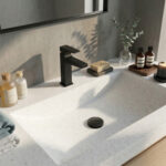 A bright, modern bathroom vanity featuring a seamless Corian Everest solid surface countertop with an integrated sloped-side sink. A matte black Hansgrohe Metropol 110 single-lever basin mixer faucet stands out against the light grey concrete-style wall. The vanity is elegantly styled with folded towels, apothecary glass bottles, a bar of soap on a dark stone dish, a lit candle, and a small glass vase holding eucalyptus branches.
