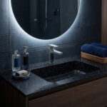 A moody, contemporary bathroom vanity showcasing a black Corian Deep Night Sky solid surface countertop with a seamless, integrated rectangular sink. A sleek, chrome Hansgrohe Metropol 110 single-lever basin mixer faucet sits centrally. The scene is softly illuminated by a large round backlit mirror against a dark grey tiled wall. Styling includes dark blue towels in a woven basket, glass dispenser bottles, a bar of soap on a marble dish, and a warm wooden vanity cabinet