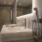 An elegant bathroom setting featuring a thick Corian White Onyx vanity top with elegant, marble-like veining. The integrated sink features a polished chrome Rebris S 110 faucet, placed next to a folded beige waffle towel and a ceramic soap dispenser. A matching chrome bidet hand shower is mounted on the textured light grey tile wall beside a backlit mirror.