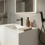 A minimalist bathroom setting featuring a seamless Corian Glacier White vanity with an integrated sink. The countertop holds a matte black Rebris S 110 faucet, an amber glass soap dispenser, and a folded beige towel. A matching black bidet hand shower is mounted on the light grey textured tiled wall next to an illuminated mirror