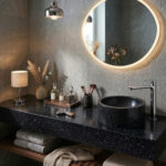 A moody and elegant bathroom setup showcasing a thick countertop and a seamless cylindrical countertop washbasin made of Corian Deep Night Sky, featuring its signature black solid surface scattered with shimmering, galaxy-like speckles. A tall, polished chrome Hansgrohe Rebris S 240 single-lever basin mixer stands next to the basin. The scene is warmly lit by a backlit round mirror, a small table lamp, and a pendant light, accompanied by glass apothecary bottles and makeup brushes on the vanity