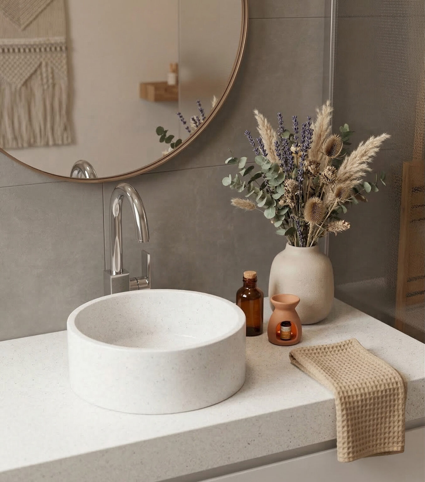 A detailed photograph of a seamlessly integrated modern bathroom vanity, featuring a light speckled Corian® Everest countertop and a matching cylindrical vessel sink with no visible seams. A polished chrome hansgrohe Vernis Shape single-lever basin mixer is elegantly installed next to the sink. To the right, a matte beige vase holds a large, dried bouquet of lavender and eucalyptus. In the background, a round mirror reflects a cozy interior with a macrame wall hanging. The setup is completed with a beige waffle-weave towel and artisan bathroom decor on the bright Corian® surface.