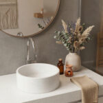 A detailed photograph of a seamlessly integrated modern bathroom vanity, featuring a light speckled Corian® Everest countertop and a matching cylindrical vessel sink with no visible seams. A polished chrome hansgrohe Vernis Shape single-lever basin mixer is elegantly installed next to the sink. To the right, a matte beige vase holds a large, dried bouquet of lavender and eucalyptus. In the background, a round mirror reflects a cozy interior with a macrame wall hanging. The setup is completed with a beige waffle-weave towel and artisan bathroom decor on the bright Corian® surface.