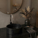 A detailed photograph of a seamlessly integrated modern bathroom vanity, featuring a Corian® Deep Night Sky countertop and a matching cylindrical vessel sink with no visible seams. A polished chrome Hansgrohe Vernis Shape single-lever basin mixer is installed next to the sink. To the right, a dark matte vase holds a luxurious arrangement of dried botanicals. In the background, a round gold-framed mirror reflects a wooden shelf against grey wall tiles. A dark towel, a wooden brush, and artisan soap rest on the dark, starry-patterned Corian® surface.