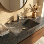 A warm, sunlit bathroom setting showcasing a chrome Hansgrohe Vivenis 110 faucet over an integrated sink on a dark, speckled Corian Lava Rock surface, styled with a vase of dry flowers.