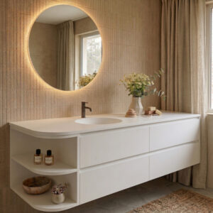A detailed view of a modern floating bathroom vanity with a smooth, seamless white Corian surface. The vanity features a centrally placed single, round vessel sink with a dark faucet. The far-left corner is designed as a multi-tiered open end shelf with rounded edges, displaying perfume bottles, a wooden bowl, and a succulent. The countertop also holds a bouquet of chamomile and eucalyptus flowers. The background shows vertically textured wall tiles, a large backlit circular mirror, a window with linen curtains, and a woven jute rug on hexagonal floor tiles.